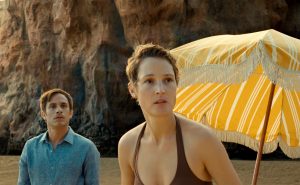 Sur la plage du film Old, Vicky Kripes en maillot de bain près d'un parasol et Gael Garcia Bernal regardent vers l'horizon, inquiets.