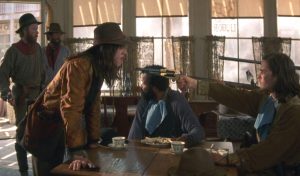 Scène d'un western réalisé par Ang Lee, où un homme, assis derrière une table, menace avec son revolver la femme cow-boy qui l'invective ; autour d'eux trois autres hommes spectateurs passifs.