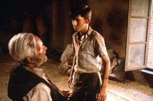 Dans la pièce vide d'une maison, un jeune adolescent se tient face à un vieillard que l'on devine ligoté et torturé sur une chaise dans le film L'échine du diable.