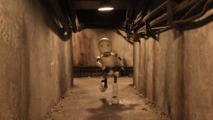 Un petit robot court dans les sous-sols d'un bâtiment industriel, avec de larges câbles au plafond, baigné dans une lumière ôcre ; scène du film Junk Head.