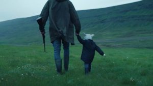 Vus de dos, une femme portant un fusil de chasse dans une main, tient un enfant de l'autre ; tous deux marchent dans une grande vallée verte ; scène du film Lamb.