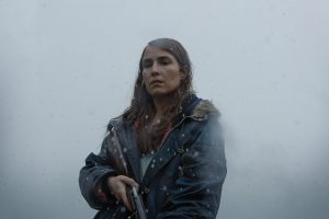 Une femme tient un fusil de chasse dans les mains, le canon baissée, au coeur d'un brouillard, le regard perdu vers le sol ; plan issu du film Lamb.