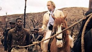 Marlon Brando sur son cheval, présente un esclave noir, une corde autour de la taille, à un potentiel acheteur dans le film Queimada.