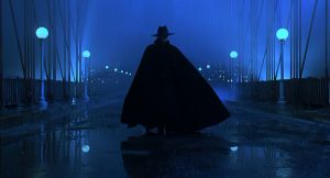 Sur un pont citadin, où le bitume humide reflète le ciel de nuit et les lampadaires bleutés, la silhouette du personnage The Shadow, avec longue cape et chapeau, vue de face.