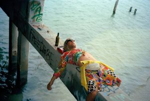 Moondog décuve sous un pont, sur une des poutres qui en font l'armature, une bouteille de bière près de sa tête ; scène du film The Beach Bum.