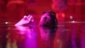 Moondog tente de fumer unecigarette dans un cours d'eau, immergé jusqu'au cou ; le tout baigné dans une irréelle luière rose et violette ; plan issu du film The beach bum.