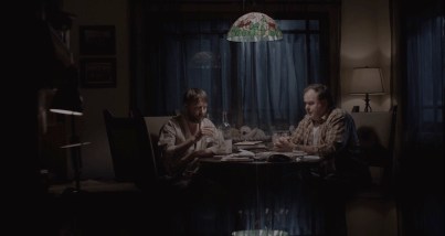 Deux hommes sont assis à la table d'un salon minable, la seule lumière vient d'un sinistre lustre au dessus de leur tête ; plan issu du film Ultrasound.