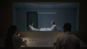 Deux individus que nous voyons de dos sont assis dans une pièce sombre, face à eux il y a une vitre qui donne sur une salle d'interrogatoire où une femme rousse questionne un accusé ; scène du film Ultrasound.