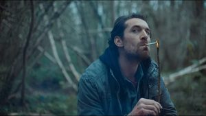 Arieh Worthalter est assis en pleine forêt ; il allume, le regard en coin, une cigarette avec une brindille de bois dans le film Hunted.