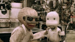 Dans une salle des machines, un scientifique avec un cerveau hypertrophié pose sa main sur l'épaule d'un petit robot triste dans le film Junk Head.