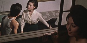 A gauche, un couple de dispute, dans l'embrasure d'une fenêtre ; à droite, à l'extérieur, l'oreille contre la vitre, une femme écoute avec malice ; scène du film La bête élagante.