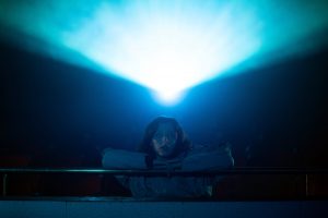Une jeune femme regarde un film juste sous le faisceau de lumière du projecteur, dans une salle de cinéma ténébreuse ; plan issu du film Al Morir la matinée.