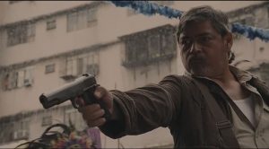 Contre-plongée sur un homme, dans la rue, qui menace quelqu'un avec un revolver ; derrière lui, des immeubles vétustes ; plan issu du film On the job 2.