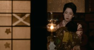 Yuri, incarnée par l'acteur masculin Bando Tamasaburo V, est chez elle, une maison dont les parois sont faites de papier fin ; elle tient une lampe à huile, l'air mélancolique ; scène du film L'étang du démon.