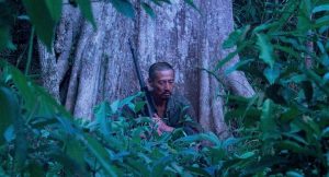 Onoda, sombre, le fusil posé à terre près de lui, médite contre un arbre dans une lueur de crépuscule ; plan du film biographique réalisé par Arthur Harari.