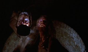 En contre-plongée, un mendiant qui a presque l'air d'un zombie tient une lampe à huile, en sous-sol ; plan issu du film Le métro de la mort.