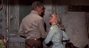 Karen Steele empêche Randolph Scott de quitter la maison, lui prenant le bras juste devant une porte en bois fermée dans le film Décision à Sundown.