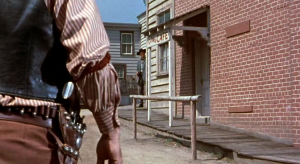 A gauche, au premier plan, le dos d'un homme qui s'apprête à dégainer en pleine rue ; face à lui à quelques mètres à droite, un homme se cache derrière une boutique ; plan issu du film Décision à Sundown.