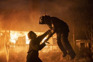 Michael Myers soulève un pompier en le plantant avec sa lame tandis qu'à l'arrière-plan, une maison brûle ; scène du film Halloween Kills.