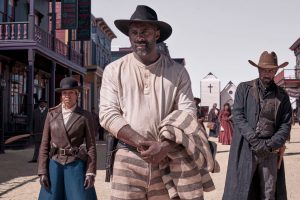 Idriss Elba en tenue de prisonnier blanche et noire, est escorté par un cow-boy à sa droite, et une femme à sa gauche, dans les rues de la ville du Far West ; scène du film The harder they fall.
