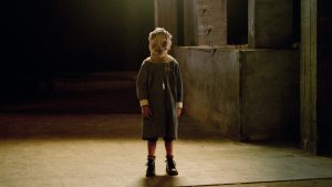 Un enfant, dans une rue sombre et de nuit, se tient droit, les bras ballants, un vieux sac en toile sur la tête, sous un lampadaire ; scène du film L'orphelinat de Juan Antonio Bayona.