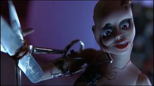 Plan rapproché-épaule sur une poupée type Barbie sans cheveux, au visage à moitié fondu et avec une petite cisaille de fortune en guise de bras gauche, dans une lumière angoissante de nuit. 