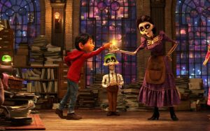 Pour notre article sur Disney 2021, une scène du film Coco, où le petit garçon du même nom, dans une bibliothèque, tend la lueur jaune qu'une femme squelette lui tend, jovialement ; derrière eux, le gardien de la bibliothèque, homme-squelette lui aussi .