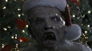 Gros plan sur le visage d'un monstre aux dents pointues et à la peau grise, portant un bonnet de Père Noël avec en fond un sapin de Noël ; issu du film Elves.