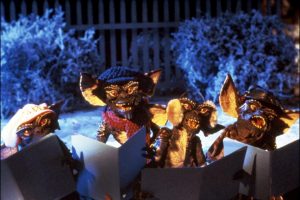 Quatre Gremlins méchants chantent des chants de Noël, lisant la partition ; ils sont dans un jardin enneigé, derrière eux, une clôture en bois.