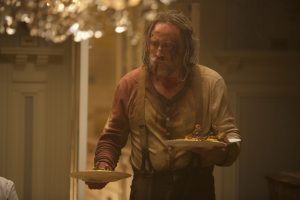 Dans une salon luxueux baigné dans une lumière jaune, Nicolas Cage abîmé, au visage et à la chemise ensanglantés, apporte deux assiettes comme  s'il était serveur d'un repas ; scène du film Pig.