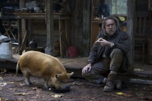Nicolas Cage assis sur le perron vieillot de sa maison en bois perdue dans la forêt, face à son cochon, Pig, qui renifle le sol.