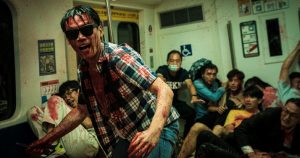 Dans une rame de métro bondée, les passagers sont tous recroquevillés les uns contre les autres tandis qu'un homme avec des lunettes de soleil exulte ; il y a du sang sur certains passagers de la rame et sur le front au premier plan ; scène du film The Sadness.