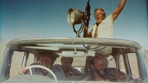 Quatre chasseurs traversent le désert en voiture, hilares, dans le film Wake in Fright.