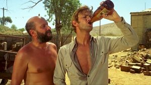 Gary Bond boit une bière au goulot, dans une rue chaude et déserte d'Australie, sous le regard amusé de Donald Pleasance dans le film Wake in Fright.