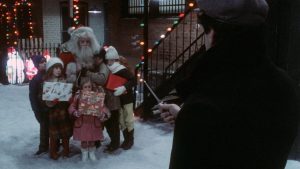 Dans une rue enneigée, un homme déguisé en Père Noël tient plusieurs enfants contre lui, pour les protéger face à un homme qui les menace d'un couteau ; scène du film Christmas Evil.
