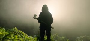 Sur fond de brume et de feuillages épais au sol, la silhouette d'un homme vu de dos, en contre-jour, portant une hache ; plan issu du film In the earth.