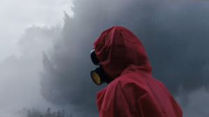 Vu de trois-quarts, un individu portant une combinaison rouge avec un masque à gaz se tient debout, en fond le brouillard gris inquiétant du film In the earth.