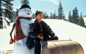 En montagne, sur un fond de neige et de sapins, une adolescente regarde l'horizon tout sourire ; derrière elle un bonhomme de neige à la mine enfantine ; scène du film Jack Frost.