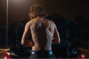 Léo, vu de dos, torse nu, range ses habits dans le coffre de sa voiture, garée sur un parking la nuit ; scène du film Hors du monde réalisé par Marc Fouchard.