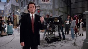 Bill Murray en costume-cravate un peu trop grand pour lui sur un plateau de tournage, semble poser une question ; derrière lui, le décor d'un film d'époque ; scène du film Fantômes en fête.