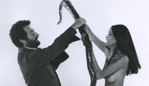 Joe d'Amato et une superbe brune seins nus face à face, avec entre eux un grand serpent, qu'ils tiennent chacun à bout de bras ; illustration pour le documentaire Inferno Rosso : Joe d'Amato.