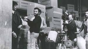 En noir et blanc, le tournage d'une scène où Joe d'Amato, cigarette au bec, montre à son comédien comment menacer avec un fusil à pompe ; dans un entrepôt rempli de cartons ; illustration pour le documentaire Inferno Rosso : Joe d'Amato.