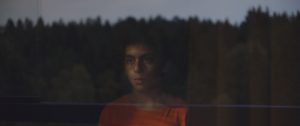 Un jeune garçon regarde un paysage de forêt, dont les arbres montent haut, à travers une fenêtre : le reflet du paysage se dessine sur la vitre et son visage ; plan issu du film The Innocents.