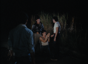 En pleine nuit, aux abords d'un champ uniquement éclairé par les phares d'une voiture, un homme apeuré est accroupi entre deux autres hommes debout ; l'un d'eux braque un revolver sur l'accroupi, prêt à tirer ; scène du film Bayan Ko.