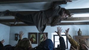 Un homme est en lévitation, plaqué, allongé, contre le plafond, au dessus de multiples individus encapuchonnés qui tendent les mains vers lui dans le film Post Mortem.