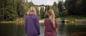 Deux jeunes filles blondes, vues de dos, observent un étang ; un immeuble se dresse, blanc, sur l'autre rive ; plan issu du film Eskil Vogt.