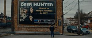 Plan d'ensemble où Michael Cimino, vu de dos, contemple une grande affiche du film Deer Hunter sur un mur en brique ; issu du documentaire Michael Cimino un mirage américain.