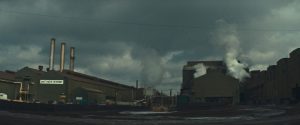 Une friche industrielle, en plan large, dont les fumées se dirigent vers un ciel gris ; scène du film Michael Cimino un mirage américain.