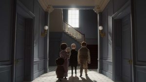 Trois individus, un homme, un garçon et une femme tenant un bébé, sont vus de dos, au bout d'un couloir juste éclairé par une petite fenêtre face à eux, au niveau d'un escalier ; scène du film La Maison.