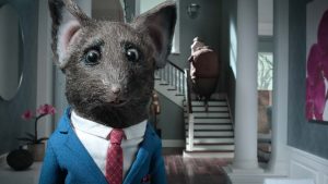 A gauche, un rat portant un costume cravate nous regarde droit dans les yeux ; à droite, à l'arrière-plan, une silhouette large vue de dos grimpe un escalier ; plan issu du film La Maison.